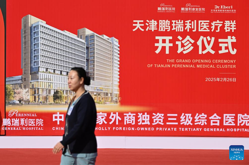 Un operatore sanitario cammina davanti a un cartellone pubblicitario per la cerimonia di apertura del Perennial General Hospital Tianjin. (26 febbraio 2025 - Xinhua/Li Ran)